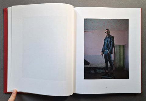 An open hardcover book with a blank white page on the left, showing a printed fashion photograph on the right page of a person in a dark outfit standing in a room. 