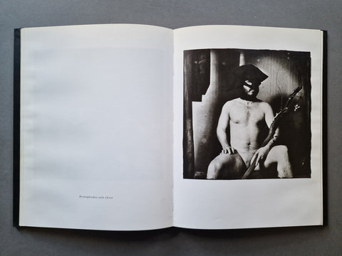 An open book showing a black-and-white photograph of a seated nude figure on the right page, with a large blank white page on the left, photographed from above on a dark gray surface.