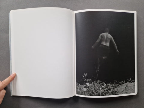 An open book or art print laid flat on a dark gray surface, showing a blank white left page and a black-and-white photographic page on the right with a silhouetted figure walking through vegetation.
