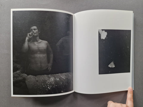 An open art book or photo magazine laid flat on a gray surface, showing two printed black-and-white photographic pages. A human finger is visible at the bottom right holding the page open.