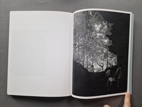 An open book or magazine lying on a dark gray surface, showing a black-and-white photographic print on the right page with a hand holding the lower edge of the page.