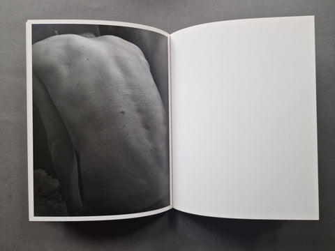 An open book or magazine laid flat on a dark gray surface, showing a black-and-white photographic print of a bare back on the left page and a blank white page on the right.