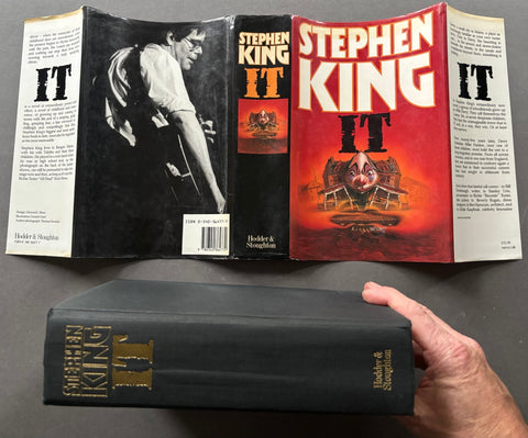 A hardcover book of Stephen King's 'IT' shown open to display the front cover, back cover, and spine, with a hand holding the black spine in the foreground against a dark gray background.