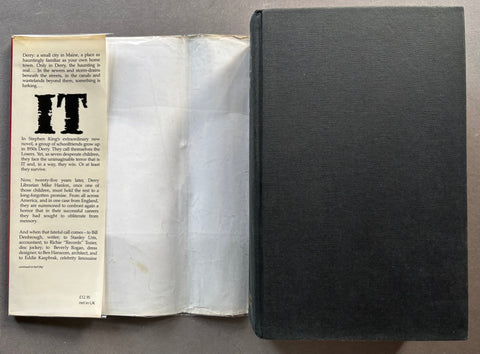 An open vintage book or booklet laid flat on a gray surface, showing a cream-colored printed interior page with a large black 'IT' heading and a dark textured black cloth-covered spine or cover standing beside it.