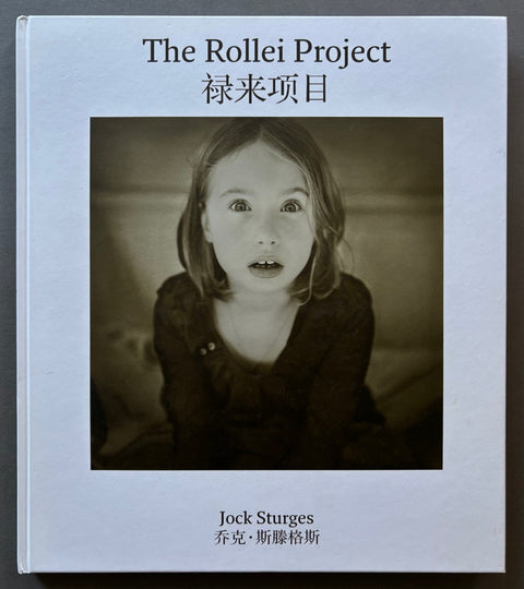 Book titled 'The Rollei Project' by Jock Sturges with a white cover and the image of a girl staring into the camera whilst her photograph is being taken.
