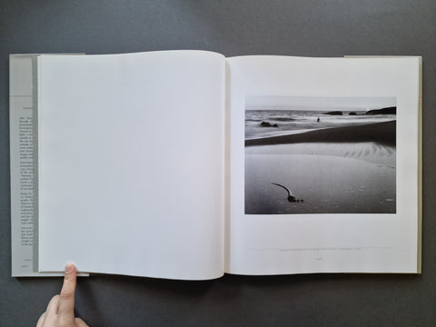 An open hardcover book: The left page is blank, while the right page features a black-and-white beach photograph with a small figure and shoreline.