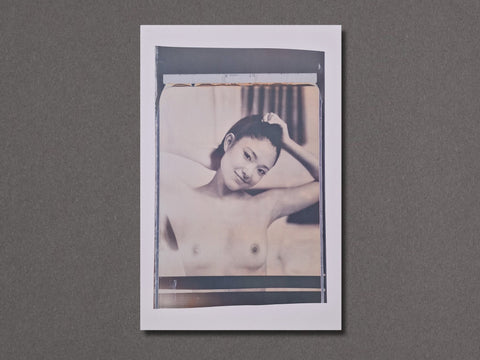 A vertical photographic art print with a white border. The photograph is a close-up of a nude woman holding her hair and smiling for the camera