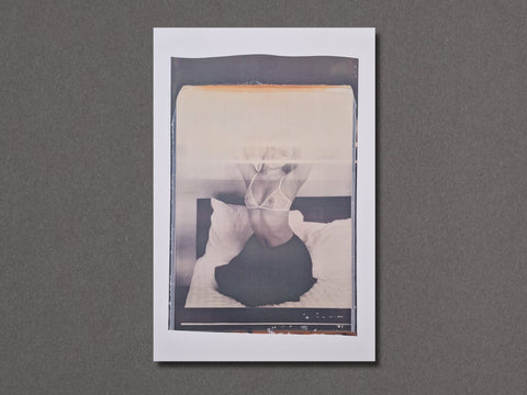 A vertical photographic art print with a white border. The photograph depicts a person sitting on a bed in a monochrome, slightly faded style with a warm beige tint at the top.