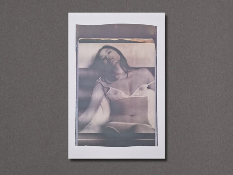 A vertical photographic art print with a white border. The photograph depicts a woman in see-through white lingerie lying on a bed