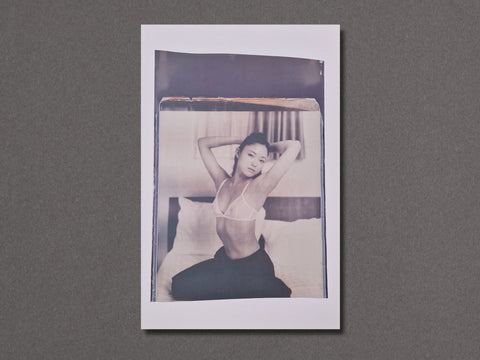A vertical photographic art print with a white border. The photograph depicts a person sitting on a bed in a monochrome, slightly faded style with a warm beige tint at the top. Close up shot