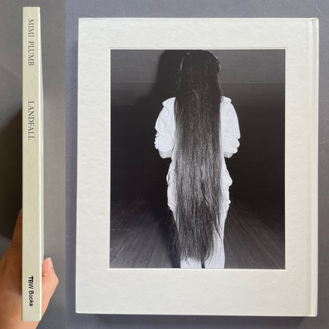 A hardcover art book or photo print shown standing upright, with the front cover featuring a black-and-white portrait of a person seen from behind with very long dark hair. The left edge shows the spine with the text 'MINI FILMFB LANDFALL' and 'TWO EYES'.