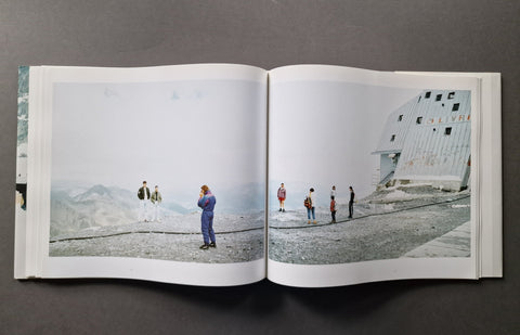 An open hardcover book, showing a large printed photograph spread across both pages. The photo depicts a snowy mountain landscape with several small people standing near a white building.