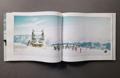 An open hardcover photo book , showing a snowy mountain landscape printed across both pages. The scene includes a large metal structure on the left page and small people on the snowy mountain on the right page.