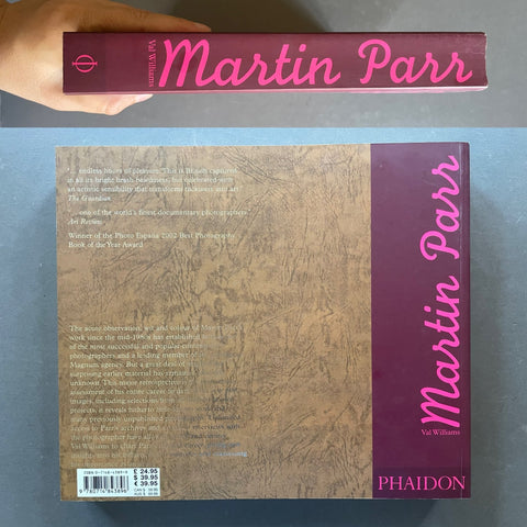 A hand holds a hardcover book titled 'Martin Parr' above the back cover of the same book on a gray surface. The cover features a textured brown background with a dark purple vertical band on the right side containing the title in pink script and the PHAIDON logo at the bottom.