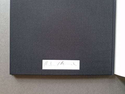 A close-up of a dark charcoal gray textured hardcover book cover with a small white rectangular label near the bottom center, photographed on a neutral gray surface.