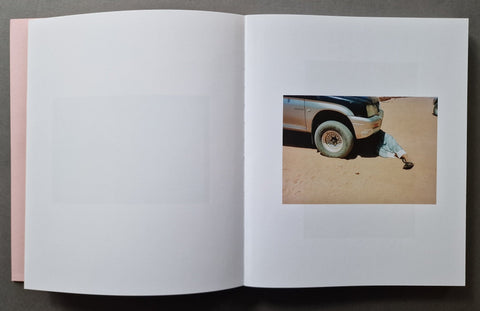 An open book , showing mostly blank white pages with a small inset photograph on the right page depicting part of a vehicle and a person on sand.