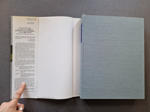 An open book with printed text on the left page and a light blue-gray fabric-covered binding on the right, photographed from above on a dark gray surface. A person's finger points to the printed text.