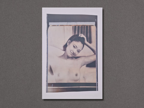 A vertical photographic art print with a white border. The photograph is a close-up of a nude woman holding her hair and smiling for the camera