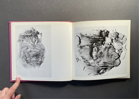 An open sketchbook or art book laid flat on a dark gray surface, showing two monochrome charcoal or pencil studies on white pages. A human finger is visible at the bottom left holding the book open.