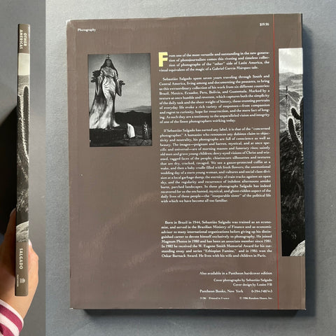 A book titled 'Other Americas' is shown lying on a gray surface, with its back cover facing up and a hand holding the spine on the left. The cover is a muted taupe-brown with black-and-white photographic imagery and a vertical red line.