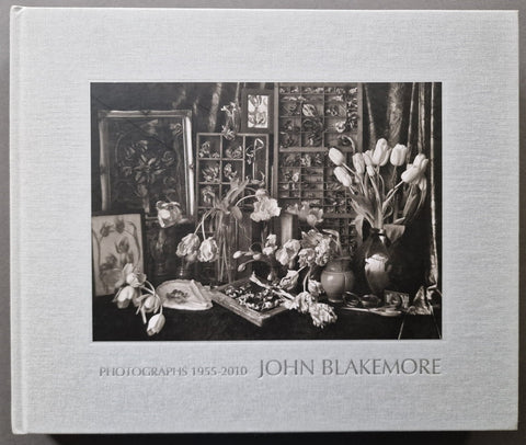 A hardcover art book titled 'Photographs 1955-2010 John Blakemore' featuring a black and white still life photograph of flowers and framed art on the cover, set against a light gray background.