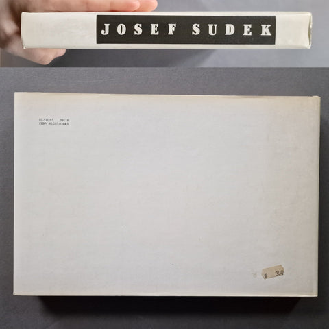 A book titled 'JOSEF SUDEK' is shown with its front cover open or laid flat below, held at the top by a hand. The cover is plain off-white with black title text and small printed text in the upper left corner.