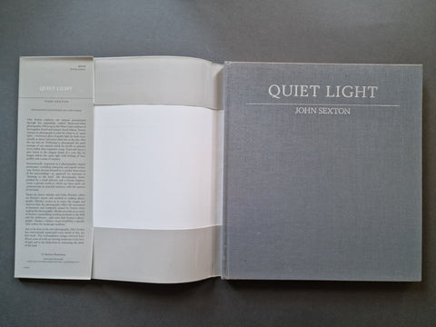 An open hardcover book titled 'QUIET LIGHT' by John Sexton, showing a gray textured cover, white interior pages, and printed text on the left page, photographed on a dark gray surface.