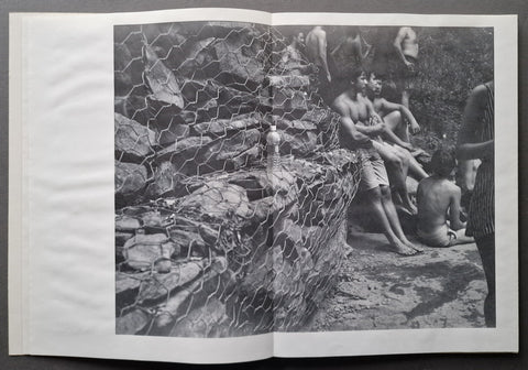 An open book spread showing a black-and-white photograph of several shirtless men sitting and standing near a rocky wall with wire mesh in the foreground.