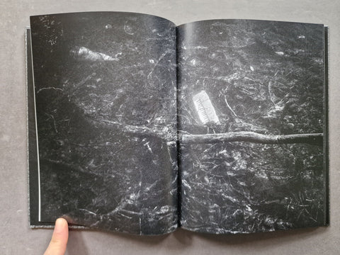 An open book showing a dark, textured black-and-white surface with a white rectangular object placed on top, photographed from above on a gray surface with a finger visible at the bottom left.