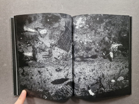 An open book showing a black and white photographic print of debris or rubble, photographed against a gray background with a finger pointing at the bottom left.