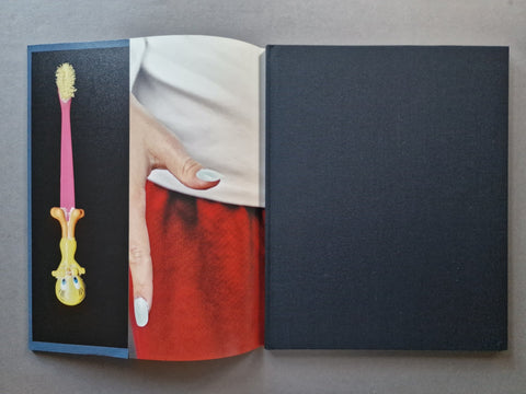 An open hardcover book - The left page features a surreal photograph with a pink and yellow novelty toothbrush on a black background next to a cropped photo of a hand with white nail polish and a red skirt.