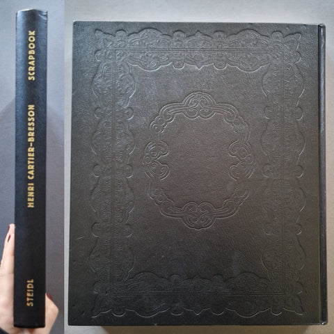 A black embossed leather-bound scrapbook book shown from the front cover and spine, held by a hand on the left. The spine reads 'SCRAPBOOK', 'HENRI CARTIER-BRESSON', and 'STEIDL' in gold lettering.