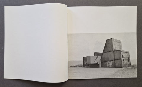 An open book, showing a black-and-white architectural photograph printed on the right page. The left page is blank, and the photo depicts a large concrete structure in an open desert landscape.