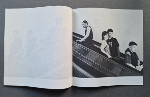 An open book, showing a black-and-white printed photograph of several people riding an escalator on the right page and a faint large monogram or logo on the left page.