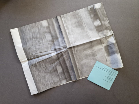 A folded square tissue paper art print with an abstract gray, white, and blue textured pattern, displayed on a dark gray surface next to a small light blue card.