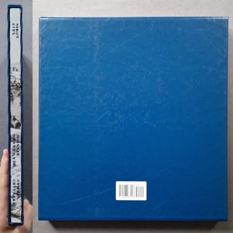 A blue hardcover book shown from the back, with a white barcode sticker near the lower center. A hand is visible holding the spine on the left for scale, against a plain gray background.
