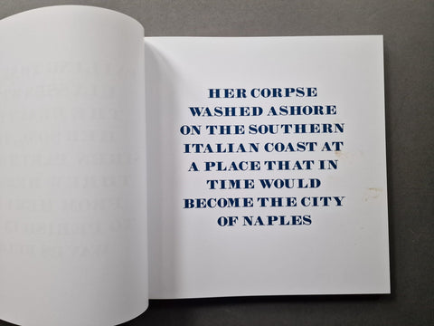 An open book showing a white page with dark blue text that reads: 'HER CORPSE WASHED ASHORE ON THE SOUTHERN ITALIAN COAST AT A PLACE THAT IN TIME WOULD BECOME THE CITY OF NAPLES.' The left page is blank or faintly visible, and the book is photographed on a dark gray surface.
