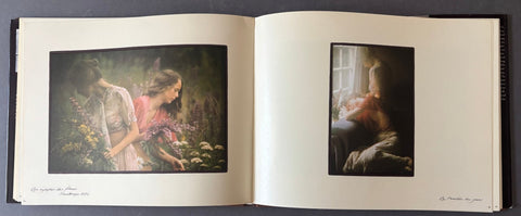 An open art book or album spread open on a dark surface, showing two framed-style printed illustrations on cream pages. The left page depicts two women among flowers in a soft, painterly scene.
