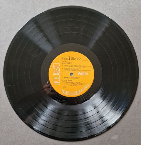 A black vinyl record of David Bowie's 'Space Oddity' on RCA Victor, featuring a bright yellow-orange label in the center, photographed flat on a gray surface.