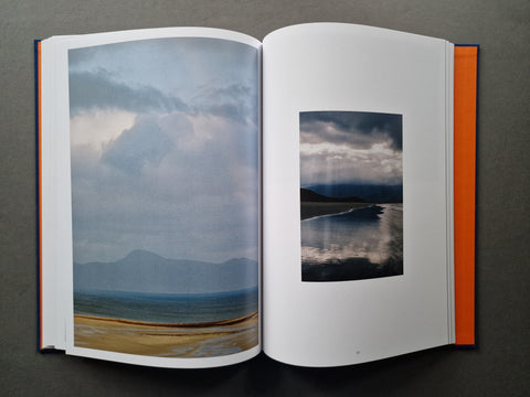 An open hardcover book lying flat on a dark gray surface, displaying two landscape photographs on its pages. The left page shows a wide coastal scene with mountains, sea, and a sandy shore under cloudy skies, while the right page features a smaller square landscape photo of a reflective shoreline.