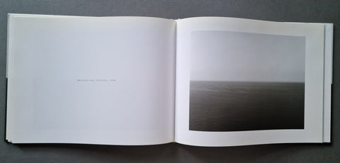 An open book showing a minimalist black and white photograph of a calm sea under a gray sky, displayed on a dark gray surface.