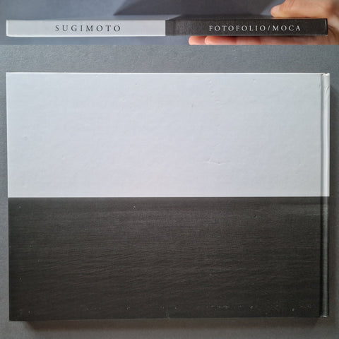 A large flat rectangular book with a white upper half and dark charcoal/black woodgrain lower half, shown on a gray surface. A smaller matching item at the top has the text 'SUGIMOTO' and 'FOTOFOLIO/MOCA', and a hand is holding it.
