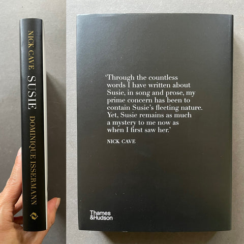 A hardcover book titled 'Susie' by Nick Cave and Dominique Issermann, shown standing upright next to a hand holding its spine. The front cover is matte black with white text containing a quote, and the spine is black with gold text.
