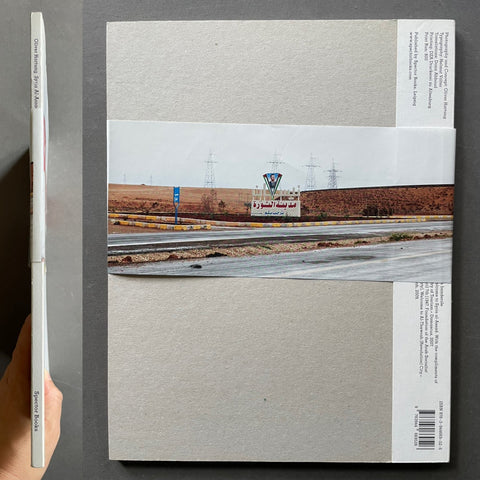 A square art print or poster with a large central landscape photograph of a highway and control tower, bordered by wide light gray textured paper sections and printed text blocks on the right and bottom edges. A hand is holding a matching slim copy of the print on the left against a dark gray background.