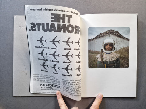 An open book spread on a gray surface, showing a black-and-white graphic page with airplane silhouettes and large upside-down text, alongside a color photograph of a person in a white space helmet. 