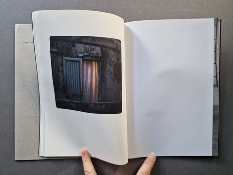 An open book on a dark gray surface, showing a printed photograph of a dark windowed wall on the left page and an empty white page on the right. 