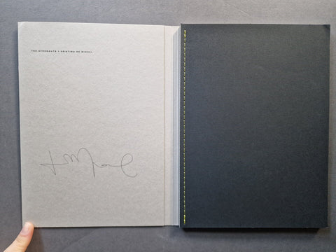 An open book - The left page is grey textured paper with small printed text near the top and a cursive signature in the lower half.