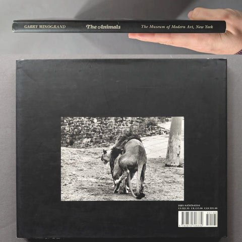 A hand holds the spine of the book 'The Animals' by Garret Winogrand, displayed above the back cover of the same book, which features a black-and-white photograph of two animals on a dark background.