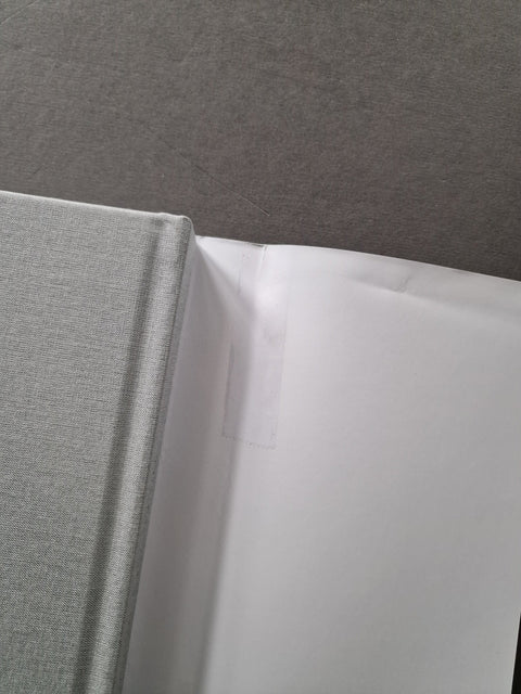 A close-up of a light gray textured hardcover book or binder cover meeting a white page or folder, set against a dark gray textured background.