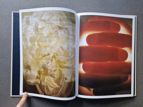 An open art book, showing two printed photographs: a large pale cream flower on the left page and a close-up of reddish-orange fingers on the right page